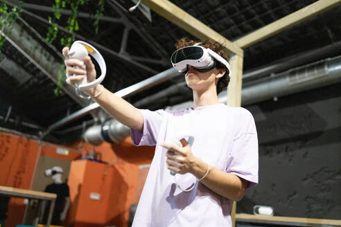 Ein junger Erwachsener, der in ein Virtual-Reality-Spiel eintaucht, ein VR-Headset trägt und die Controller fokussiert und konzentriert hält. - ADSF50480