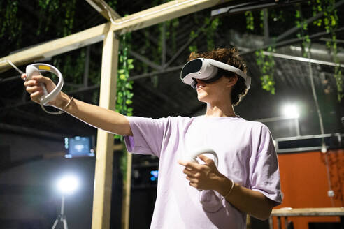 Ein fröhlicher junger Mensch spielt mit einem Virtual-Reality-Headset und Controllern unter der Abendbeleuchtung, völlig eingetaucht in eine virtuelle Welt. - ADSF50483