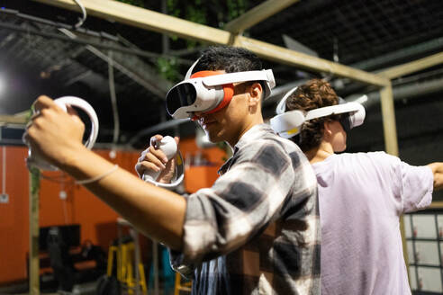 Side view of two friends stand in a gaming area fully immersed in a virtual reality world wearing VR headsets and holding motion controllers - ADSF50491