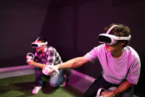 Two friends stand in a gaming area fully immersed in a virtual reality world wearing VR headsets and holding motion controllers in neon light room - ADSF50492