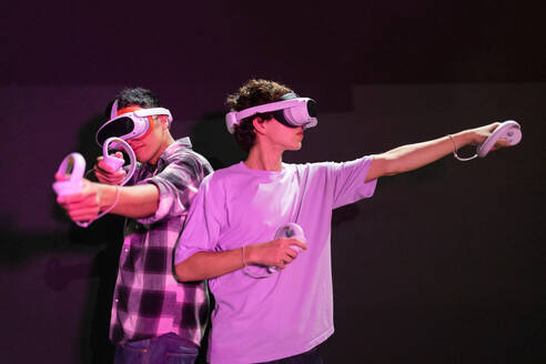 Two friends stand in a gaming area fully immersed in a virtual reality world wearing VR headsets and holding motion controllers in neon light room - ADSF50493