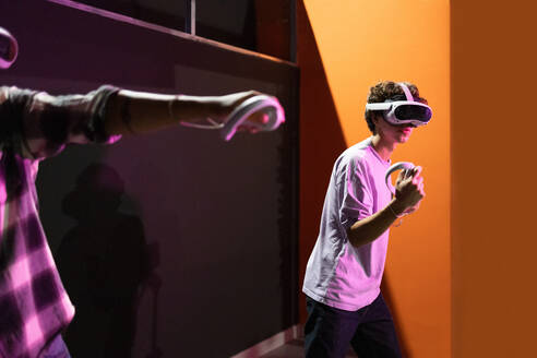Seitenansicht von zwei Freunden, die in einem Spielbereich stehen und vollständig in eine Welt der virtuellen Realität eintauchen, VR-Headsets tragen und Bewegungssteuerungen in einem Raum mit Neonlicht halten - ADSF50494
