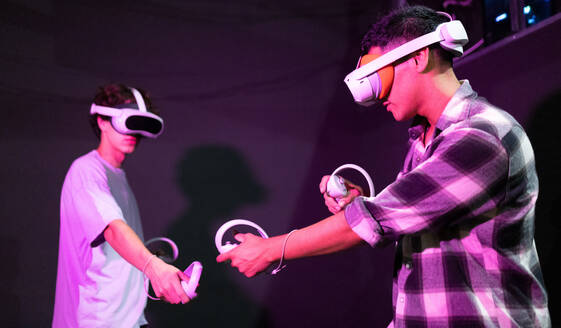 Side view of two friends stand in a gaming area fully immersed in a virtual reality world wearing VR headsets and holding motion controllers in neon light room - ADSF50495