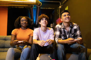 Drei junge, gemischtrassige Freunde sitzen auf einer Couch und spielen ein lustiges Videospiel - ADSF50506