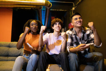 Drei junge, multirassische, glückliche, siegreiche Freunde, die auf einer Couch sitzen und mit geschlossener Faust feiern, während sie ein lustiges Videospiel spielen - ADSF50507
