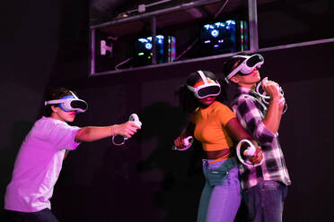 Aufgeregte, gemischtrassige Freunde, die ein Virtual-Reality-Spiel genießen, jeder mit VR-Headset und Controller in der Hand, eingetaucht in ein digitales Abenteuer - ADSF50517