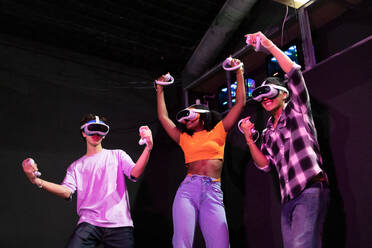 Aufgeregte multirassische Freunde, die siegessicher durch ein Virtual-Reality-Spiel hüpfen, jeder mit VR-Headset und Controller in der Hand, eingetaucht in ein digitales Abenteuer - ADSF50519