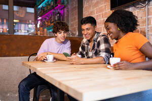 Fröhliche multiethnische Gruppe von Freunden mit Tassen Kaffee in einem gemütlichen Café, die auf ihrem Mobiltelefon surfen - ADSF50523