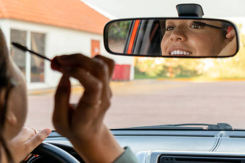 Lächelnde glamouröse junge kaukasische Geschäftsfrau, die in den Rückspiegel schaut und Wimperntusche aufträgt, während sie tagsüber in einem modernen Auto sitzt - ADSF50532