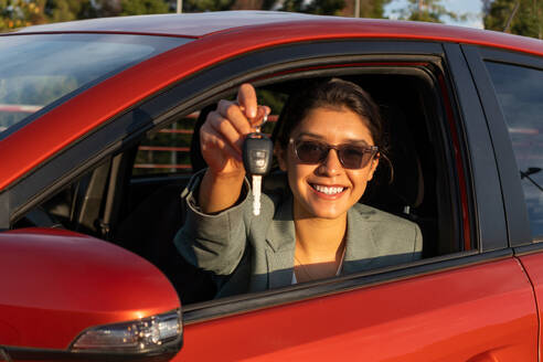 Porträt einer fröhlichen kaukasischen schönen jungen Verkäuferin mit Sonnenbrille, die lächelt und den Schlüssel zeigt, während sie in einem modernen orangefarbenen Auto bei Sonnenuntergang sitzt - ADSF50533
