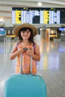 Portrait of cute girl wearing casuals and straw hat holding blue suitcase and standing against arrival timetable while waiting for departure at airport - ADSF50538