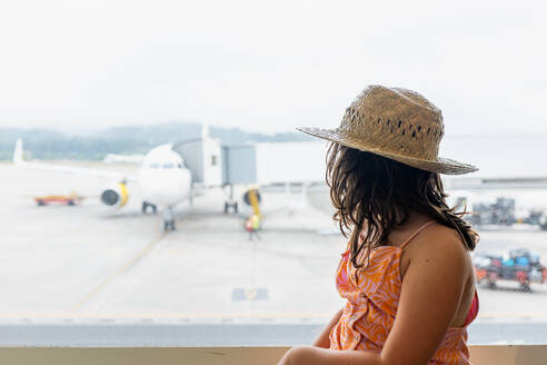 Seitenansicht einer gesichtslosen Reisenden mit Strohhut und Freizeitkleidung, die ein Passagierflugzeug auf der Rollbahn durch ein Fenster am Flughafen betrachtet - ADSF50543