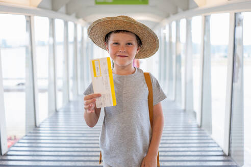Porträt eines niedlichen lächelnden Jungen in Freizeitkleidung und Strohhut, der an der Fluggastbrücke steht und seine Bordkarte im Flughafenterminal zeigt - ADSF50544