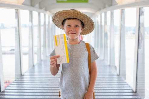 Portrait of cute smiling boy in casuals and straw hat standing at boarding bridge and showing boarding pass in airport terminal - ADSF50544