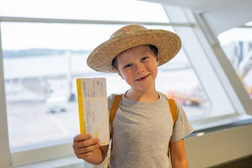 Porträt eines niedlichen lächelnden Jungen in Freizeitkleidung und Strohhut, der an der Fluggastbrücke steht und seine Bordkarte im Flughafenterminal zeigt - ADSF50545