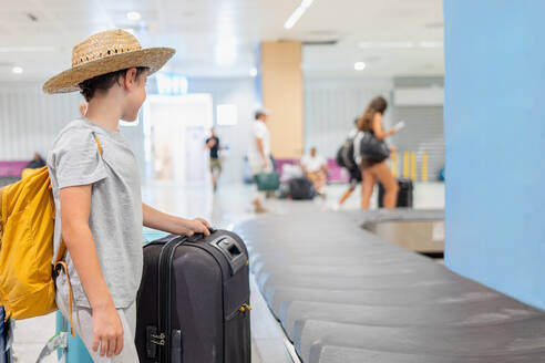 Seitenansicht eines anonymen Jungen in Freizeitkleidung und Strohhut mit Rucksack und Koffer, der am Förderband steht und auf sein Gepäck am Flughafen wartet - ADSF50549