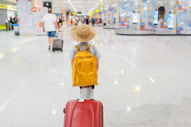 Rückenansicht eines anonymen Kindes mit Hut und Rucksack, das einen roten Koffer zieht, während es durch ein beleuchtetes modernes Terminal läuft - ADSF50550