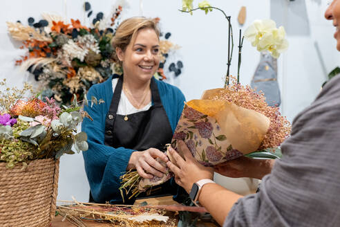 Glücklich lächelnde Blumenhändlerin, die einem Kunden in einem Blumenladen, umgeben von verschiedenen Blumen, einen Blumenstrauß verkauft - ADSF50551