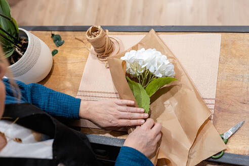 Draufsicht auf die Hände einer nicht erkennbaren Person, die einen Blumenstrauß bastelt und weiße Blumen in braunes Papier auf einem Holztisch einwickelt, mit Garn und Schere in der Nähe - ADSF50553