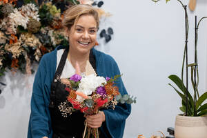 Eine Floristin lächelt in die Kamera, während sie in ihrem Geschäft einen schönen Blumenstrauß arrangiert, mit einer stilvollen Blumenkulisse - ADSF50554