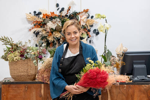 Lächelnde Floristin, die in einem Blumenladen steht und in die Kamera schaut, hält einen leuchtend roten Blumenstrauß in der Hand, umgeben von einer Vielzahl bunter Blumenarrangements in einer Werkstatt - ADSF50556