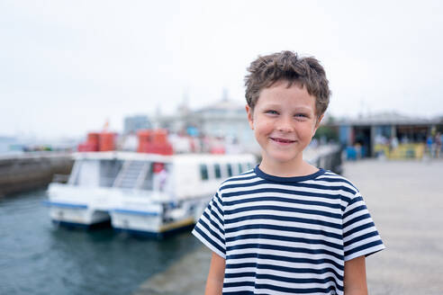 Fröhliches Kind im gestreiften T-Shirt lächelt in die Kamera mit dem Meer und Booten im Hintergrund während der Sommerferien - ADSF50558