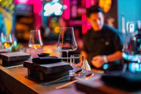 Essstäbchen mit gefalteter schwarzer Serviette und leeren Trinkgläsern auf einem ordentlichen Holztisch an einer modernen beleuchteten Sushi-Bar vor einem verschwommenen männlichen Koch in schwarzer Uniform - ADSF50562