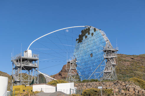 Blick auf das Observatorium des Roque de los Muchachos auf der Palmeninsel: Die komplizierte Struktur des Observatoriums hebt sich von einem klaren blauen Himmel ab, während der Mond bei Tageslicht ruhig im Hintergrund zu sehen ist und von Grünflächen umgeben ist - ADSF50566