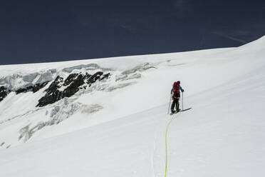 Back view of anonymous male backpacker with skies and poles walking on snow covered mountain against blue sky at Swiss Alps on sunny day - ADSF50577