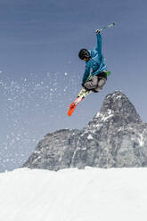 Ganzer Körper eines unerkennbaren aktiven Mannes, der nach unten schaut, während er mit Skiern und Skischläger über verschneite Berggipfel gegen den blauen Himmel im Urlaub in den Schweizer Alpen springt - ADSF50580