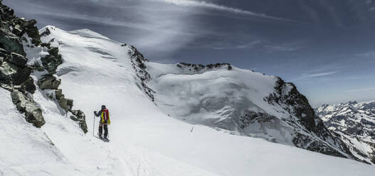 Rückenansicht einer anonymen Person mit Rucksack und Stöcken, die auf schönen verschneiten Bergen gegen den dunklen Himmel wandert, während sie ihren Winterurlaub in den Schweizer Alpen genießt - ADSF50582