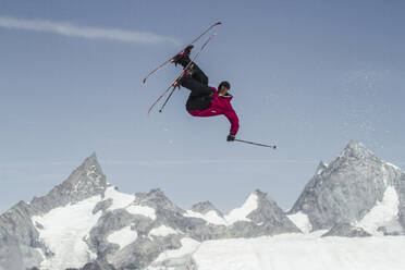 Full body of carefree active male skier dressed in warm clothes with skies and poles jumping above snow covered mountain while enjoying winter vacation at Swiss Alps - ADSF50585