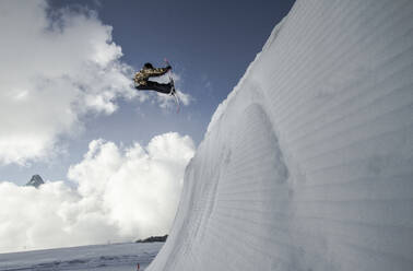 Side view of anonymous man in warm clothes jumping with snowboard above snowy mountain against blue cloudy sky during winter vacation at Swiss Alps - ADSF50593