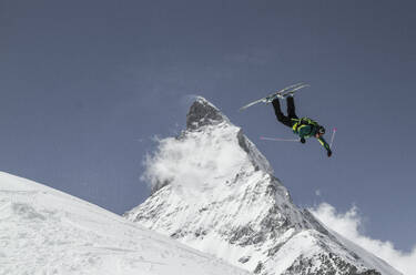 Full body of carefree active male skier dressed in warm clothes with skies and poles jumping above snow covered mountain while enjoying winter vacation at Swiss Alps - ADSF50600