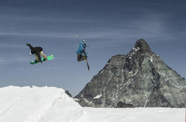 Ganzkörperansicht eines anonymen männlichen Freundes mit Snowboard und Skiern, der über eine schneebedeckte Landschaft mit blauem Himmel in den Schweizer Alpen springt - ADSF50616