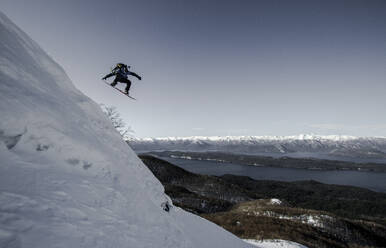 Full body of anonymous person in warm clothes jumping with snowboard above snow covered mountain slope against beautiful mountains and clear sky during vacation - ADSF50618