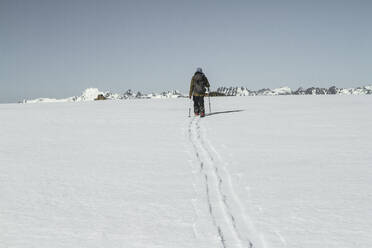 Back view full body of unrecognizable person with backpack skiing on snow covered landscape under clear blue sky during sunny day during winter vacation - ADSF50630