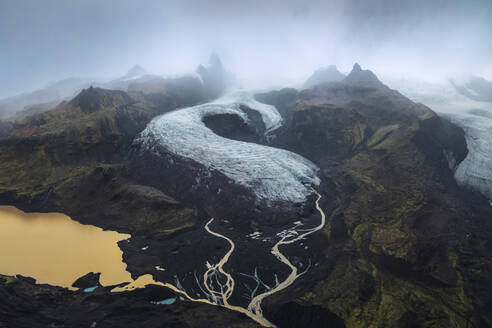 Fesselnde Luftaufnahme eines Gletschers, der sich durch den Vatnajokull-Nationalpark in Island schlängelt, umgeben von zerklüftetem Terrain und bewölktem Himmel. - ADSF50692