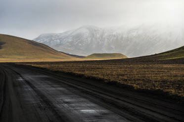 Eine ruhige Szene im weiten Hochland von Island, die eine unbefestigte Straße zeigt, die sich sanft durch die karge Landschaft vor schneebedeckten Bergen unter einem sanft beleuchteten Himmel windet - ADSF50702