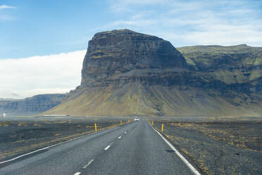 Ein ruhiger Blick auf eine offene Straße, die zu einem massiven, zerklüfteten Berg im einsamen isländischen Hochland führt und die karge Schönheit und Größe des Geländes verdeutlicht - ADSF50709