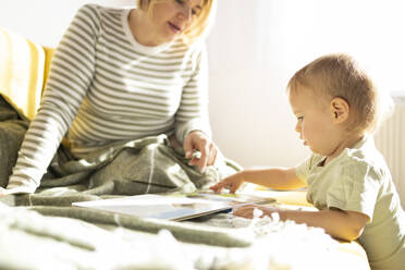 Kleinkind mit einem Buch neben einer wachsamen Mutter in einem sonnenbeschienenen Zimmer. - ADSF50711