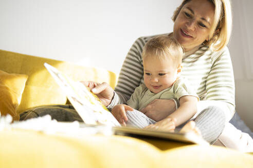 Eine lächelnde Mutter liest ihrem aufmerksamen Kleinkind aus einem Bilderbuch vor, während sie auf einer gemütlichen gelben Couch sitzt. - ADSF50713