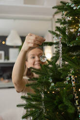 Ein fröhliches junges Mädchen hängt konzentriert funkelnde Ornamente an die üppigen Zweige eines Weihnachtsbaums - ADSF50715