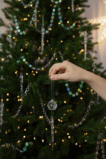 Die Hand einer Person hängt ein zartes Ornament an einen festlich geschmückten Weihnachtsbaum. - ADSF50716
