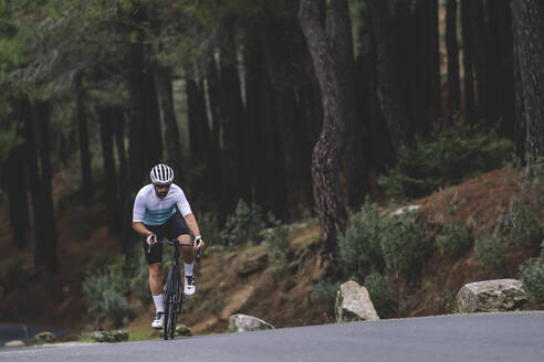 Cyclist riding uphill on a forested mountain road dressed in a white and teal jersey - ADSF50717