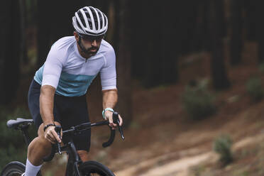 Konzentrierter Radfahrer in weißem und blaugrünem Trikot, der eine Kurve auf einem Waldweg fährt - ADSF50719