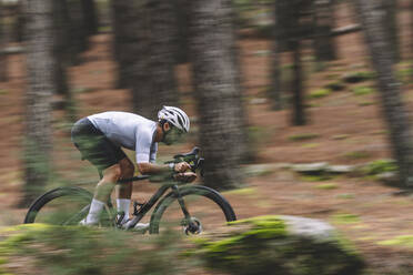 Radfahrer in Bewegung in einem weißen und blaugrünen Trikot, der durch einen Kiefernwald fährt - ADSF50720