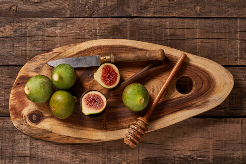 Overhead shot of a rustic wooden cutting board with fresh green figs, a half-cut fig, a honey dipper, and an old knife on a wooden background. - ADSF50724