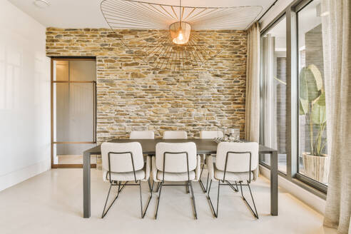 Stilvolles Licht über Esstisch und Stühlen im Esszimmer mit Backsteinwand und großem Fenster in einem modernen Haus - ADSF50733