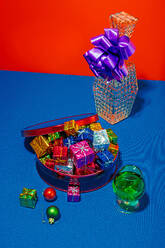 Von oben bunte Weihnachtsgeschenke in einer roten Dose und eine glitzernde Vase mit einer lila Schleife auf einem leuchtend blauen Tisch vor rotem Hintergrund - ADSF50762